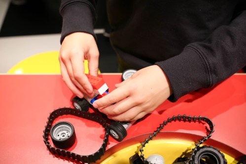 A child’s hands assembling a toy vehicle with wheels and tracks on a colorful table.