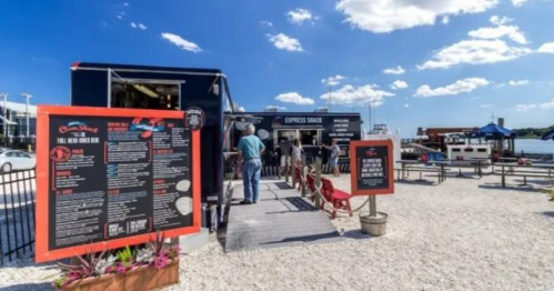 A food truck by the water with a menu board, people in line, and a sunny sky with scattered clouds.