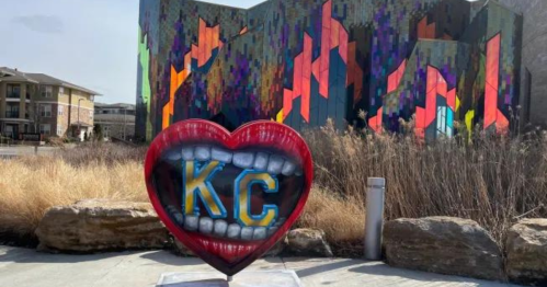 A colorful heart sculpture with "KC" in its mouth, set against a vibrant, patterned building and dry grass.