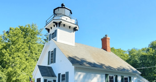 A white lighthouse with a black lantern sits atop a building surrounded by trees under a clear blue sky.
