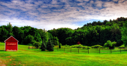 A vibrant green landscape featuring a red barn, trees, and a wooden fence under a partly cloudy sky.