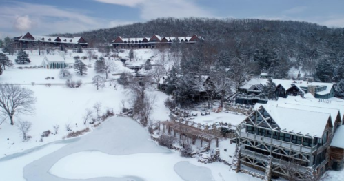 A snowy landscape featuring a lodge and surrounding trees, with a frozen pond in the foreground and hills in the background.