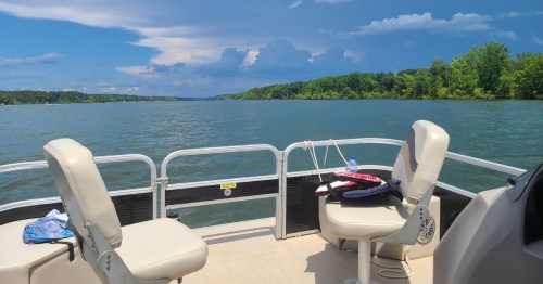 View from a boat on a calm lake, with green trees lining the shore and a blue sky filled with clouds.