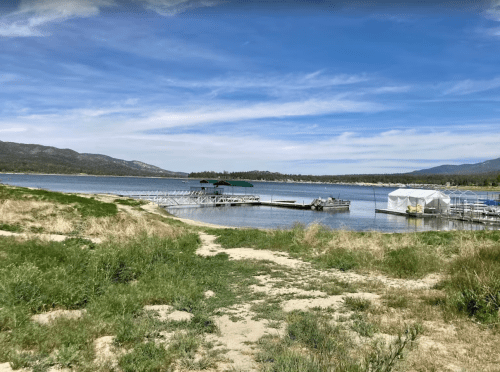 A serene lakeside view with docks, boats, and grassy shores under a blue sky with scattered clouds.