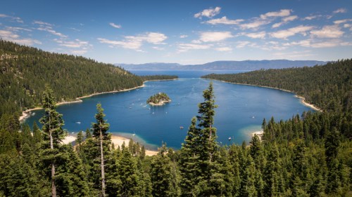 A scenic view of a tranquil lake surrounded by lush green forests and distant mountains under a clear blue sky.