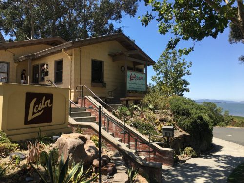 A charming yellow building with a sign reading "Leila" near a waterfront, surrounded by greenery and steps leading up.