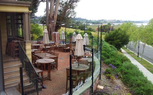 Outdoor dining area with wooden tables and umbrellas, overlooking a scenic landscape and water in the background.