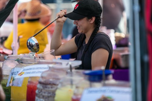 A woman in a black cap serves drinks from colorful jars at a market stall, smiling as she uses a ladle.