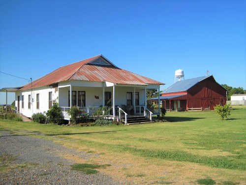 A white house with a rusty roof and a red barn in a grassy field under a clear blue sky.