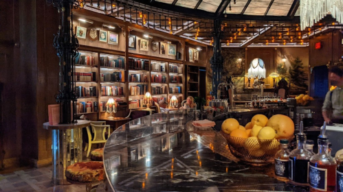 A cozy bar with a marble counter, bookshelves, warm lighting, and a bowl of oranges in a stylish setting.