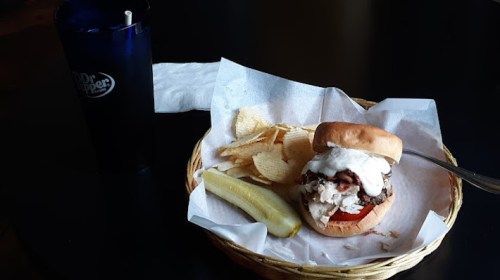 A burger with toppings, potato chips, a pickle, and a Dr Pepper drink in a blue cup on a table.