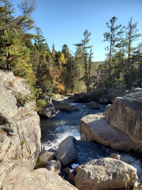 A serene river flows between large rocks, surrounded by trees with autumn foliage under a clear blue sky.