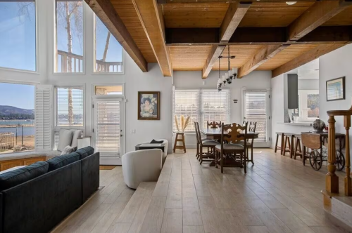 Bright, open living space with wooden beams, large windows, and a dining area overlooking a lake. Modern and inviting decor.
