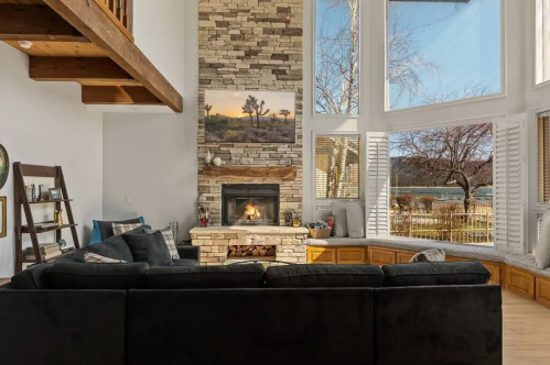 Cozy living room with a stone fireplace, large windows, and a view of nature, featuring a dark sectional sofa.