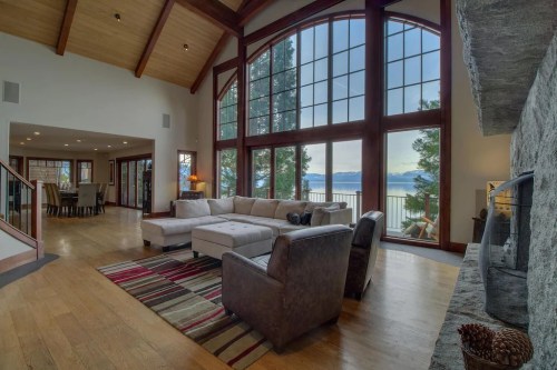 Spacious living room with large windows, lake view, cozy seating, and a stone fireplace. Bright and inviting atmosphere.