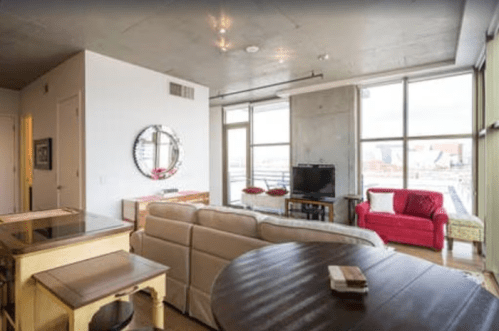 Modern living room with large windows, a beige sofa, red armchair, wooden dining table, and a TV on a stand.