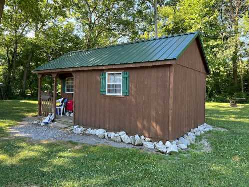 A small brown cabin with a green metal roof, surrounded by grass and trees, featuring a porch and stone border.