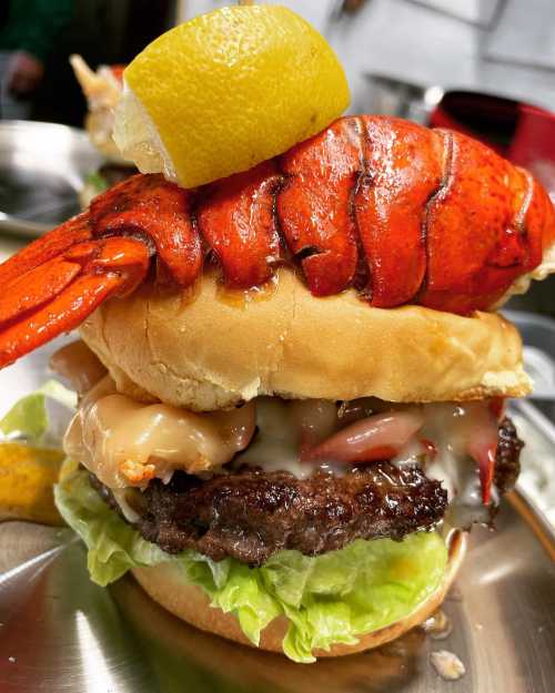 A towering burger with a beef patty, lettuce, and a whole lobster claw topped with a lemon slice.