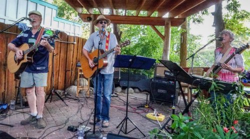 Three musicians perform on stage outdoors, playing guitars and keyboards, surrounded by greenery and a wooden structure.