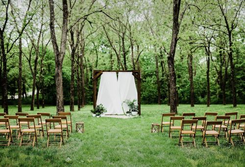 Outdoor wedding setup with wooden chairs facing a draped altar surrounded by lush green trees.