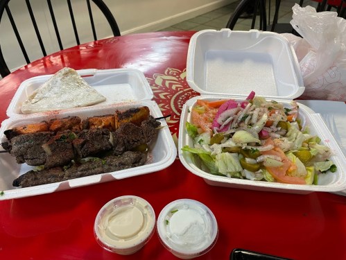 Two takeout containers on a red table: one with skewers and flatbread, the other with a fresh salad and sauces.