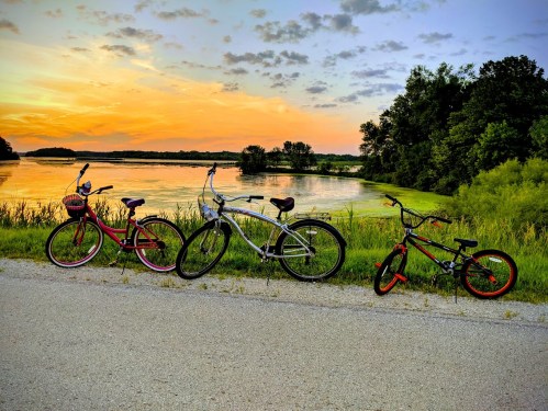 Three bicycles parked by a serene lake at sunset, with vibrant colors in the sky and lush greenery surrounding the water.