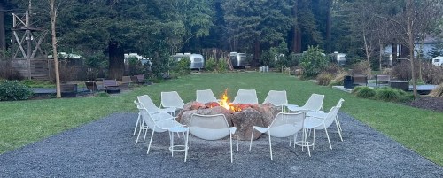 A circular seating area with white chairs around a fire pit, surrounded by greenery and airstream trailers in the background.