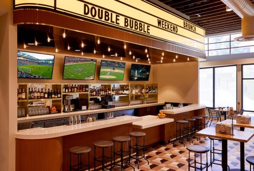 A modern bar with a sleek design, featuring multiple screens, a well-stocked shelf, and seating for patrons.