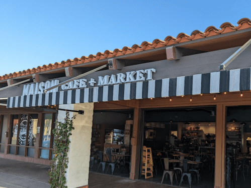 Exterior of Maison Café + Market with a striped awning and outdoor seating under a clear blue sky.