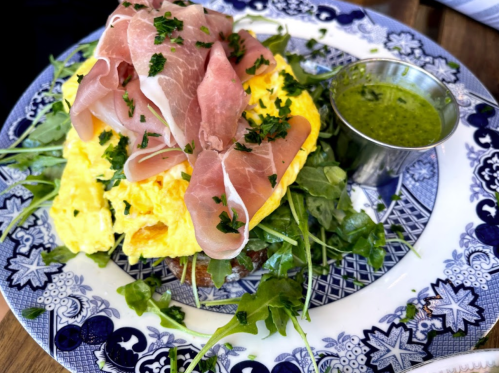 A colorful plate featuring an omelet topped with prosciutto, arugula, and a small cup of green sauce.