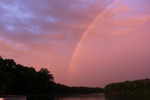 A serene river scene at sunset, featuring a vibrant rainbow arching over lush green trees and calm waters.