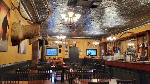 Cozy restaurant interior with wooden tables, antler decor, and a vintage ceiling, featuring a bar and warm lighting.