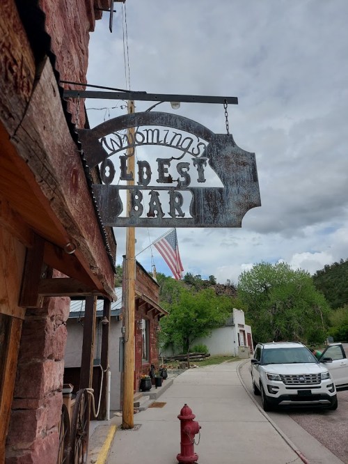 Sign for "Wyoming's Oldest Bar" with an American flag in the background, set against a rustic building and cloudy sky.