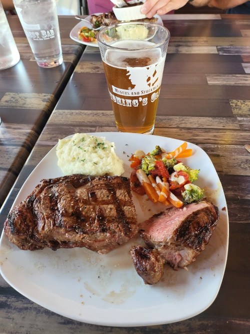 A plate with grilled steak, mashed potatoes, and mixed vegetables, alongside a glass of beer on a wooden table.