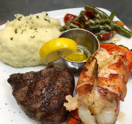 A plate featuring grilled lobster tail, steak, mashed potatoes, and sautéed green beans with a lemon wedge.