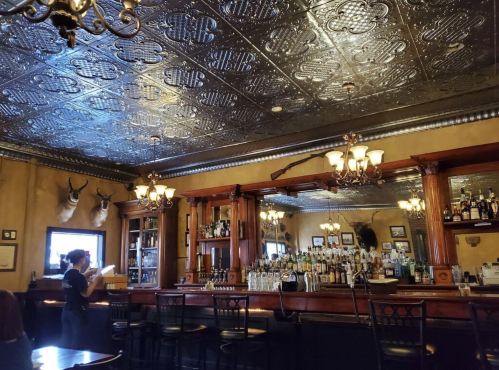 A vintage bar interior with a mirrored back, ornate ceiling, and taxidermy decor, featuring a bartender serving drinks.