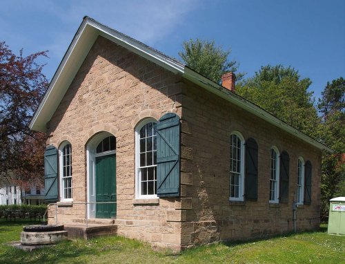 A historic stone building with green shutters and a front door, surrounded by trees and grass under a clear blue sky.