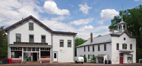 A white, two-story general store with a bell tower building nearby, set against a blue sky with clouds.