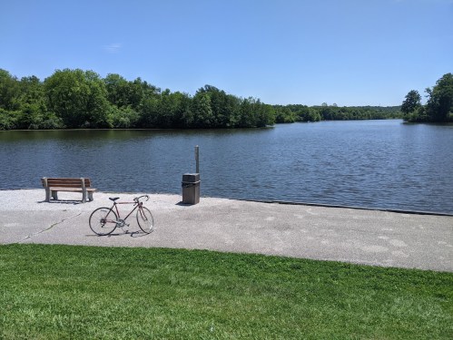 A serene lakeside view with a bike parked on a path, surrounded by green trees and a clear blue sky.