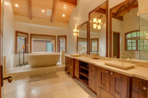 Spacious bathroom with a freestanding tub, double sinks, wooden cabinetry, and large mirrors under a vaulted ceiling.