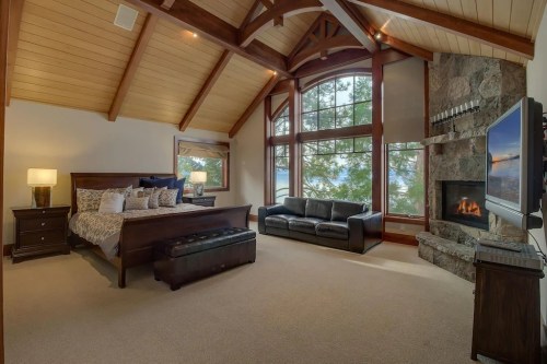 Cozy bedroom with a large window, wooden beams, a fireplace, and a seating area, overlooking a scenic view.