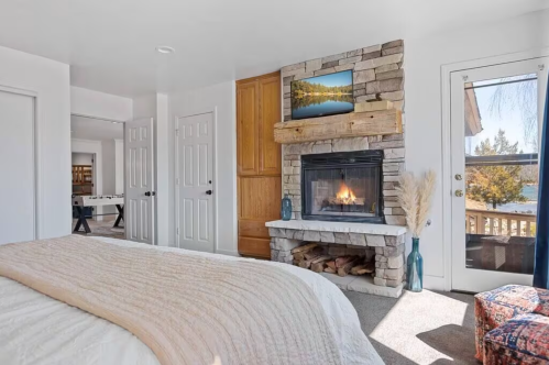 Cozy bedroom with a stone fireplace, TV above, and large windows showcasing a scenic view. Warm, inviting decor.