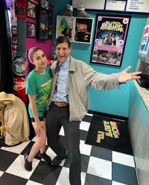 A man and a woman pose playfully in a colorful retro-themed room with movie posters and a checkered floor.