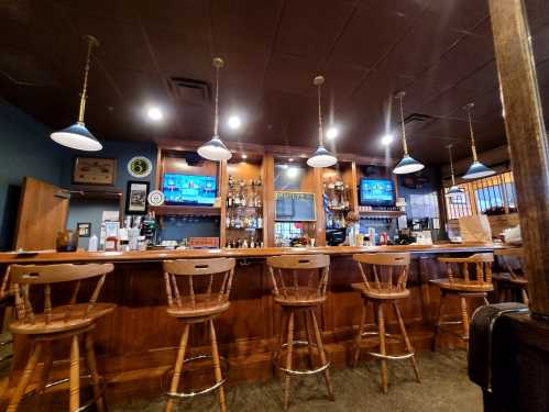 A cozy bar with wooden stools, shelves of liquor, and two TVs displaying sports in a warmly lit setting.