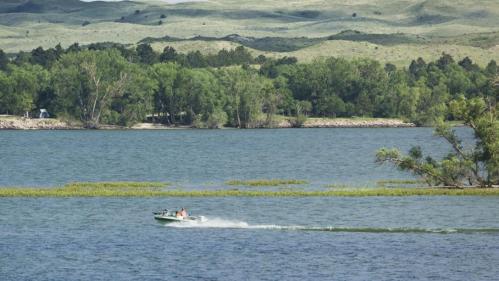A small boat speeds across a calm lake, surrounded by lush greenery and rolling hills in the background.