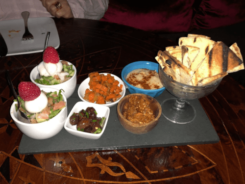 A platter featuring various appetizers: salads, roasted carrots, olives, dip, and toasted bread, garnished with raspberries.