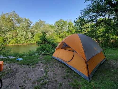 A bright orange tent set up by a river, surrounded by lush greenery and clear blue skies.