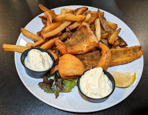 A plate of crispy fried fish fillets and golden fries, served with two dipping sauces and a lemon wedge.