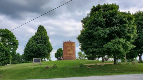 A large "Welcome" sign at a camping resort, surrounded by green trees and a cloudy sky.