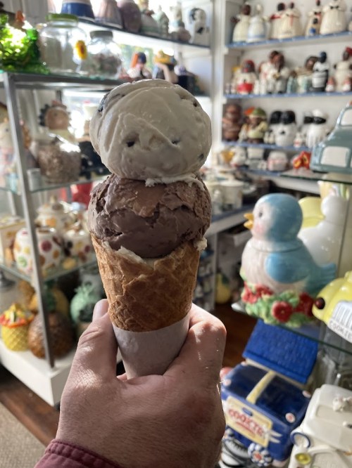 A hand holding a cone with two scoops of ice cream, surrounded by colorful ceramic collectibles in a shop.